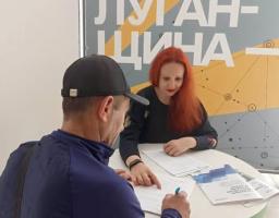 За круглим столом чоловік у кепці сидить спиною до камери та заповнює документи під наглядом жінки з яскравим рудим волоссям, а на задньому плані стоїть великий інформаційний банер із великим написом «ЛУГАНЩИНА»