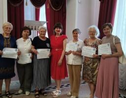 Сім літніх жінок стоять у ряд у світлій кімнаті з бордовими шторами, тримаючи в руках сертифікати, при цьому жінка по центру одягнена в яскраву червону сукню.