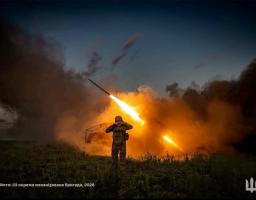 Український військовослужбовець стоїть у полі на тлі густого диму та закриває вуха руками в момент яскравого вогняного залпу реактивної артилерії у вечірніх сутінках.