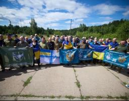 Велика група українських військовослужбовців та звільнених захисників позує на відкритій місцевості під синім небом, тримаючи в руках державні прапори України та різні військові стяги, зокрема 36-ї окремої бригади морської піхоти.