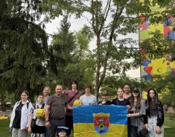 Група дорослих та дітей позує для спільного фото на вулиці біля зелених дерев, тримаючи синьо-жовтий прапор із гербом Новоайдара та кілька жовто-блакитних футбольних м'ячів.