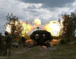 На зображенні зафіксовано момент потужного вогняного спалаху під час пострілу замаскованої артилерійської установки серед дерев, за яким зліва спостерігають двоє військових у камуфляжі