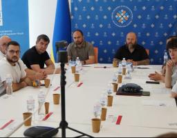 Група чоловіків сидить за великим білим Т-подібним столом під час офіційного засідання на тлі брендованих синіх банерів ПРООН та Конгресу місцевих та регіональних влад