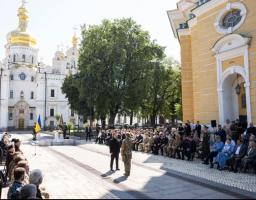 На світлині зображено офіційну церемонію на подвір'ї Києво-Печерської лаври, де Президент України Володимир Зеленський вручає державну нагороду військовому перед численними запрошеними гостями та військовослужбовцями на тлі величних білокам'яних храмів із золотими банями.