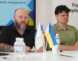 На зображенні двоє чоловіків сидять за столом під час офіційної зустрічі чи презентації, де ліворуч сидить бородатий чоловік із татуюваннями на руках у чорній сорочці, праворуч — чоловік у зеленій футболці-поло, а на передньому плані перед ними стоять маленькі прапорці України та інші настільні прапори поруч із пляшками води.
