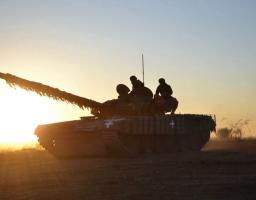 Силует українського танка із замаскованим стволом та екіпажем на броні рухається полем на тлі яскравого вечірнього або ранкового сонця, що заходить за горизонт