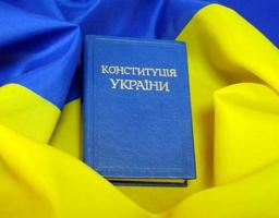 На зображенні представлена синя книга з тисненим золотим написом «КОНСТИТУЦІЯ УКРАЇНИ», яка лежить у центрі на об'ємних складках синьо-жовтого державного прапора.