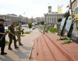 На світлині зображено трьох чоловіків в уніформах (поліцейського, військового та службовця), які з білими квітами в руках підходять Алеєю Героїв Небесної Сотні у Києві до меморіальних плит із портретами загиблих для вшанування їхньої пам'яті.