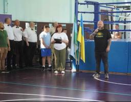 Чоловік із мікрофоном виступає перед групою людей біля боксерського рингу, де також встановлено державний прапор України.