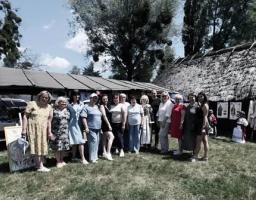Група людей стоїть у ряд на траві біля дерев'яних торговельних яток та традиційної будівлі зі стріхою під час ярмарку просто неба в оточенні дерев.