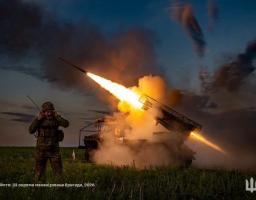 На зображенні показано вечірній запуск ракети з української реактивної системи залпового вогню, де військовослужбовець на передньому плані закриває вуха руками від гучного звуку пострілу