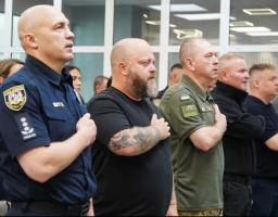Ряд чоловіків у військовій, поліцейській та цивільній формі стоять у приміщенні та тримають праву руку на серці під час урочистої події або виконання гімну.