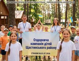 На світлині у лісовому таборі група усміхнених дітей та двоє вожатих тримають великий банер «Оздоровча кампанія дітей Щастинського району станом на 01.07.2026», тримаючи в руках маленькі прапорці України на тлі дерев'яних будиночків