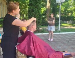 На світлині просто неба у парку жінка-перукар стриже чоловіка, який сидить у колісному кріслі, загорнутий у бордову накидку-пеньюар, тоді як на задньому плані за ними спостерігає інша усміхнена жінка.
