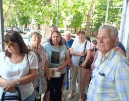 Група людей середнього та літнього віку стоїть у черзі під навісом будівлі на тлі зелених дерев вулиці.