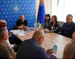 На фотографії зображено робочу зустріч групи людей за довгим дерев'яним столом у кабінеті на тлі синього банера з логотипом Конгресу місцевих та регіональних влад і прапорів, де учасники обговорюють документи, використовують ноутбук та мобільний телефон.