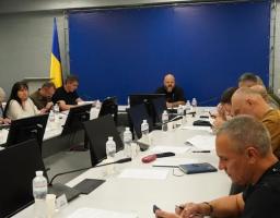 Офіційне засідання групи людей за великим П-подібним столом із моніторами, де в центрі на тлі синього банера сидить лисий бородатий чоловік, а ліворуч у кутку стоїть прапор України.