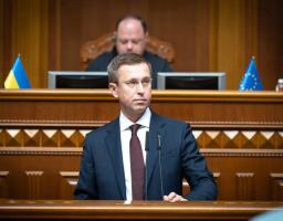 Світлина із сесійної зали Верховної Ради України, де за трибуною з мікрофонами стоїть чоловік у темно-синьому костюмі та окулярах, а на задньому плані за дерев'яною президією з прапорами України та ЄС сидить інший чоловік.