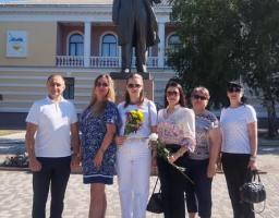 Група з шести людей у літньому повсякденному одязі, деякі з яких тримають квіти, стоїть на вимощеній площі та позує для спільного фото перед високим бронзовим пам'ятником чоловікові у костюмі на тлі двоповерхової адміністративної будівлі з колонами.