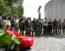 Група людей у цивільному одязі та військовій формі стоїть у шерензі на брущатці просто неба, тримаючи в руках синьо-жовті букети квітів під час офіційного заходу вшанування пам'яті, на тлі дерев, високої колони та сірої бетонної будівлі.