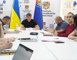 У світлому залі за великим білим столом із паперами та гаджетами проходять робочі збори, де в центрі на тлі прапорів та символіки Луганської обласної військової адміністрації сидить спікер із табличкою «Іван БОЧКО», а з обох боків від нього розмістилися інші учасники та учасниці зустрічі.