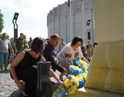 Люди, серед яких жінка у вишиванці, присіли біля підніжжя меморіалу на брущатці, щоб покласти великі синьо-жовті букети квітів під час офіційної церемонії вшанування пам'яті на тлі круглої сірої будівлі та інших учасників заходу.