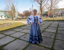 На світлині посеред скверу стоїть усміхнена жінка у вишиванці та довгій блакитній спідниці з прозорим верхнім шаром, тримаючи руки на талії