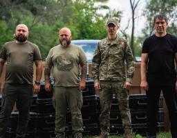 На тлі зелених дерев та коробок стоять четверо чоловіків у темному одязі та військовій камуфляжній формі, троє з яких дивляться прямо в кадр, а один зліва злегка повернув голову.