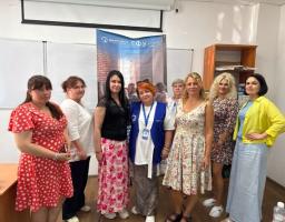 У світлій аудиторії на тлі білої дошки та вертикального банера з логотипом «ГФУ» стоять вісім жінок у різноколірному літньому одязі, вишикувавшись у ряд та дивлячись у бік камери