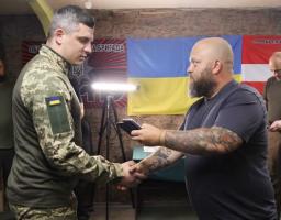 У приміщенні на тлі українського прапора та військової символіки татуйований чоловік у темній футболці тисне руку військовослужбовцю у камуфляжній формі та передає йому смартфон, поки двоє інших чоловіків спостерігають за процесом на задньому плані.