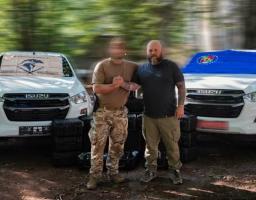 На ґрунтовій дорозі між двома білими пікапами Isuzu з прапорами на лобовому склі стоять двоє чоловіків — ліворуч військовий у камуфляжних штанах із замиленим обличчям, а праворуч бородатий чоловік у чорній футболці, які міцно тиснуть один одному руки на тлі складених на землі чорних кейсів із технікою.