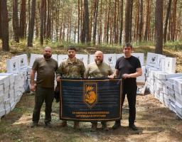 У сосновому лісі на тлі чисельних стосів білих картонних коробок стоять четверо чоловіків у військовому та цивільному одязі, які тримають перед собою великий чорний прапор із символікою та написом «60 ОКРЕМА МЕХАНІЗОВАНА ІНГУЛЕЦЬКА БРИГАДА».
